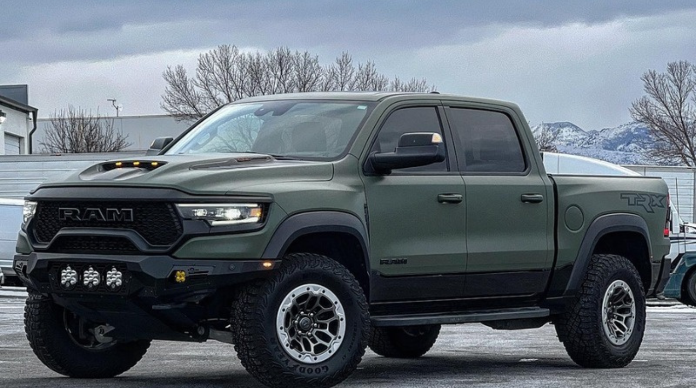 A matte green RAM TRX pickup truck, a true luxury car, is parked outdoors on pavement, with mountains and trees visible in the background under a cloudy sky.