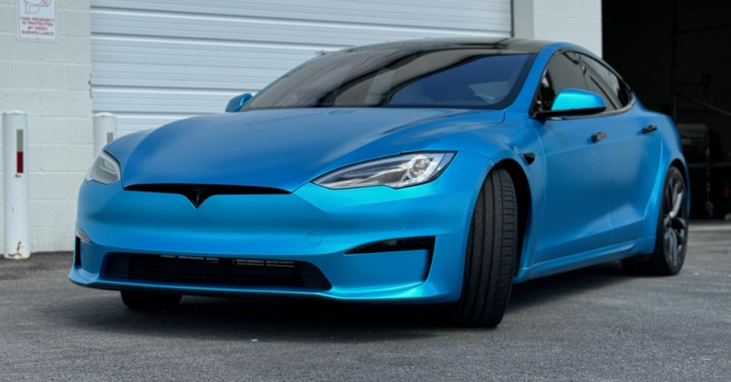 A blue Tesla sedan with sleek vehicle wraps is parked in front of a white garage door on a paved surface.