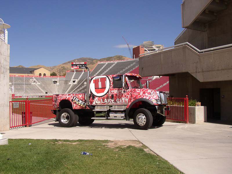 A large red and white truck featuring “U” and “Salt Lake City” graphics is parked near a stadium entrance with concrete stands and a mountain in the background, showcasing striking fleet graphics.