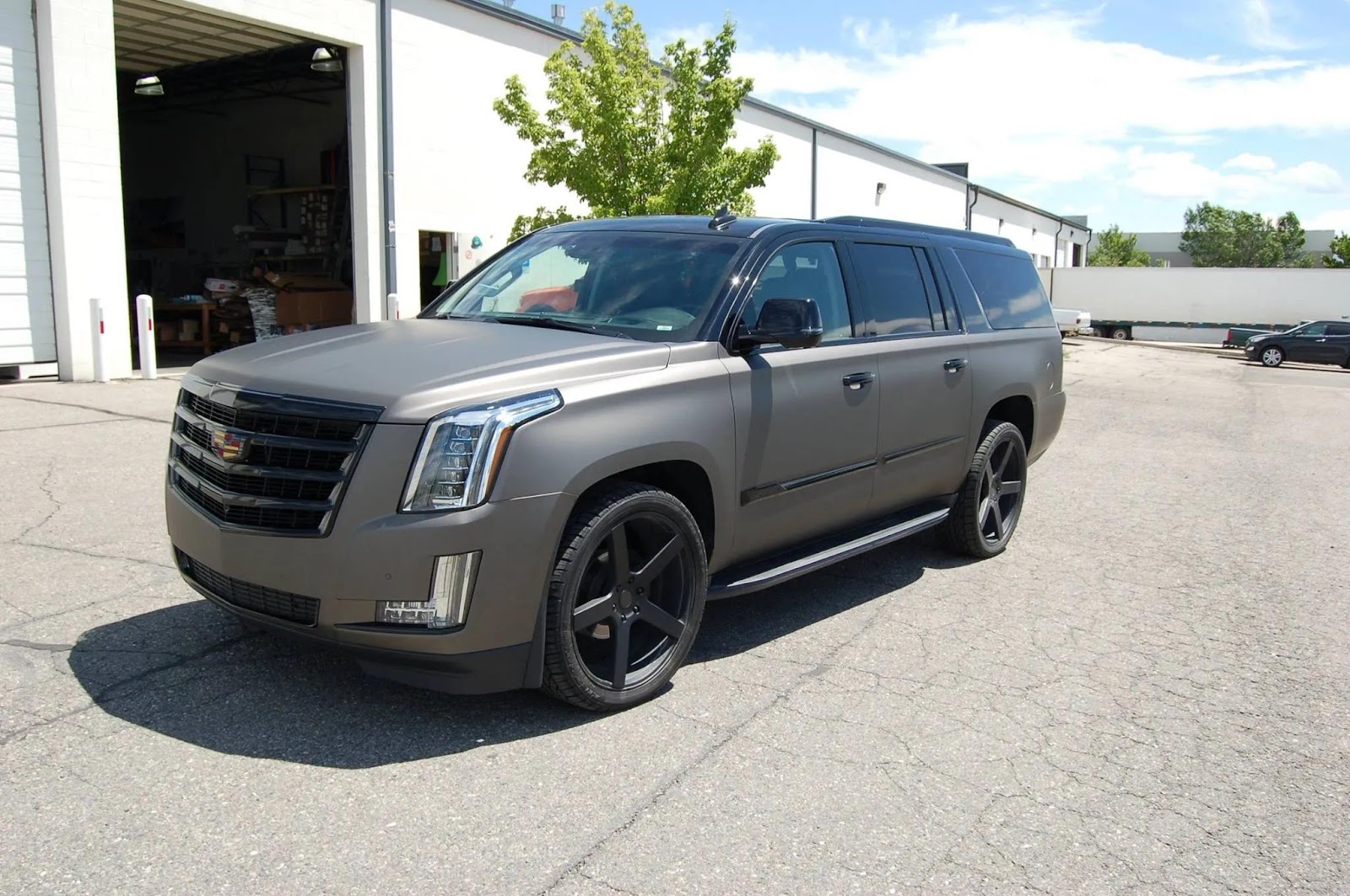 A matte gray Cadillac Escalade SUV with black wheels, featuring sleek window tinting, is parked outside an industrial building on a sunny day.