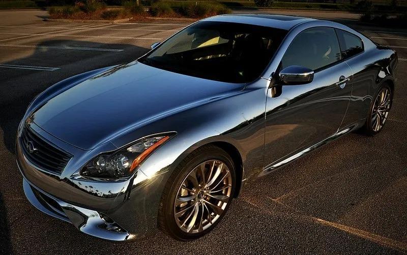 A chrome-wrapped sports coupe with expertly applied window tinting is parked on an asphalt lot under sunlight, reflecting its surroundings on its shiny exterior.