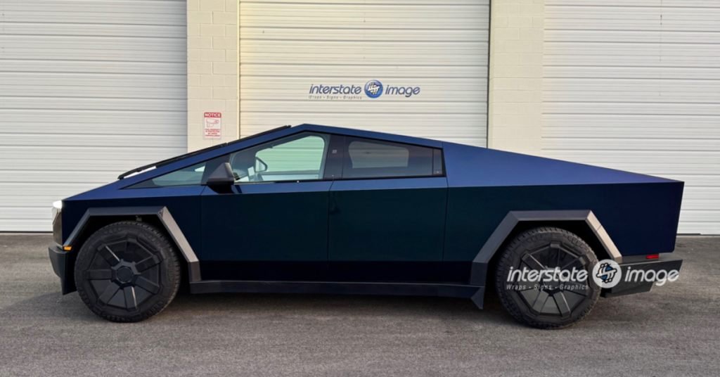 A matte dark blue Tesla Cybertruck with a 3M Certified vehicle wrap is parked in front of a white garage door featuring "interstate image" branding, showcasing top-notch business branding solutions.