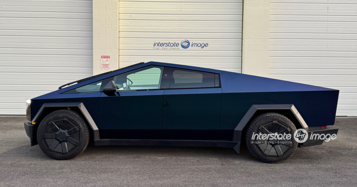 A matte dark blue Tesla Cybertruck with a 3M Certified vehicle wrap is parked in front of a white garage door featuring "interstate image" branding, showcasing top-notch business branding solutions.