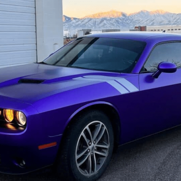 A purple Dodge Challenger with custom vehicle stripes parked in front of a white building, snow-capped mountains in the background—capturing the bold spirit of Salt Lake City.