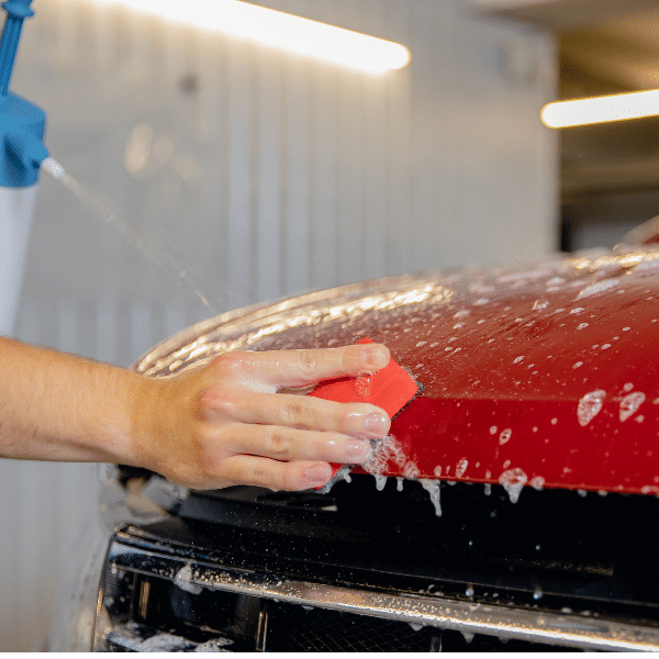 A person washes the hood of a red car with a red sponge, using soap and water in an indoor setting, maintaining car wrap durability for maximum protection.