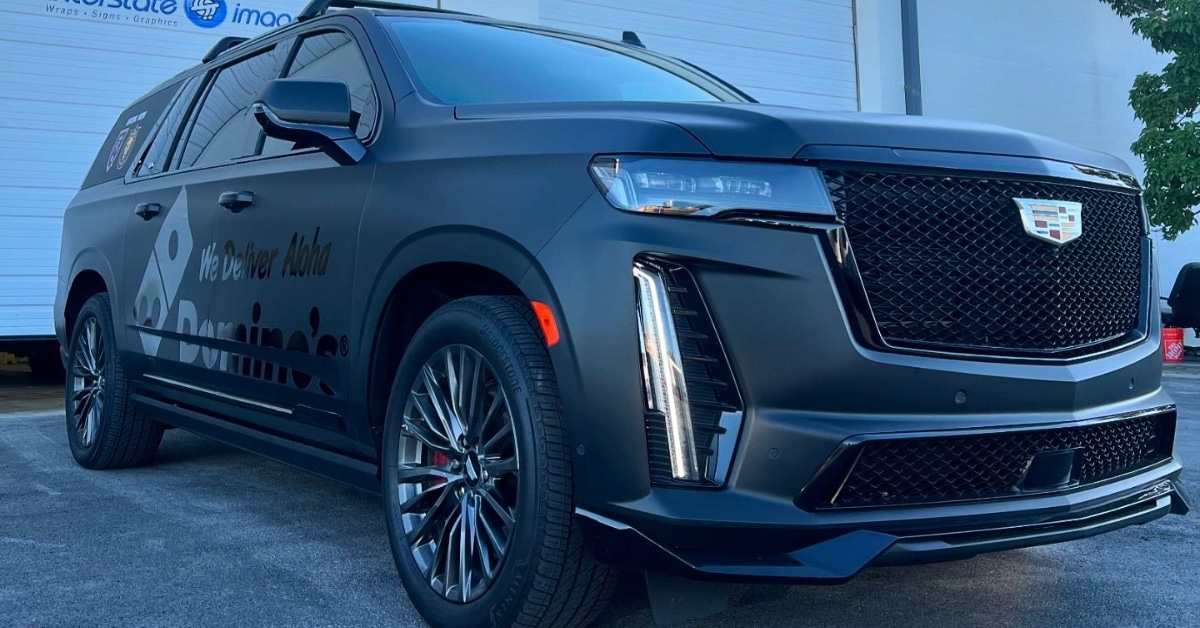 A matte black Cadillac SUV, enhanced with a color change wrap, is parked in front of a building. Its tinted windows and chrome accents add to its sleek appearance, showcasing the cost of luxury.